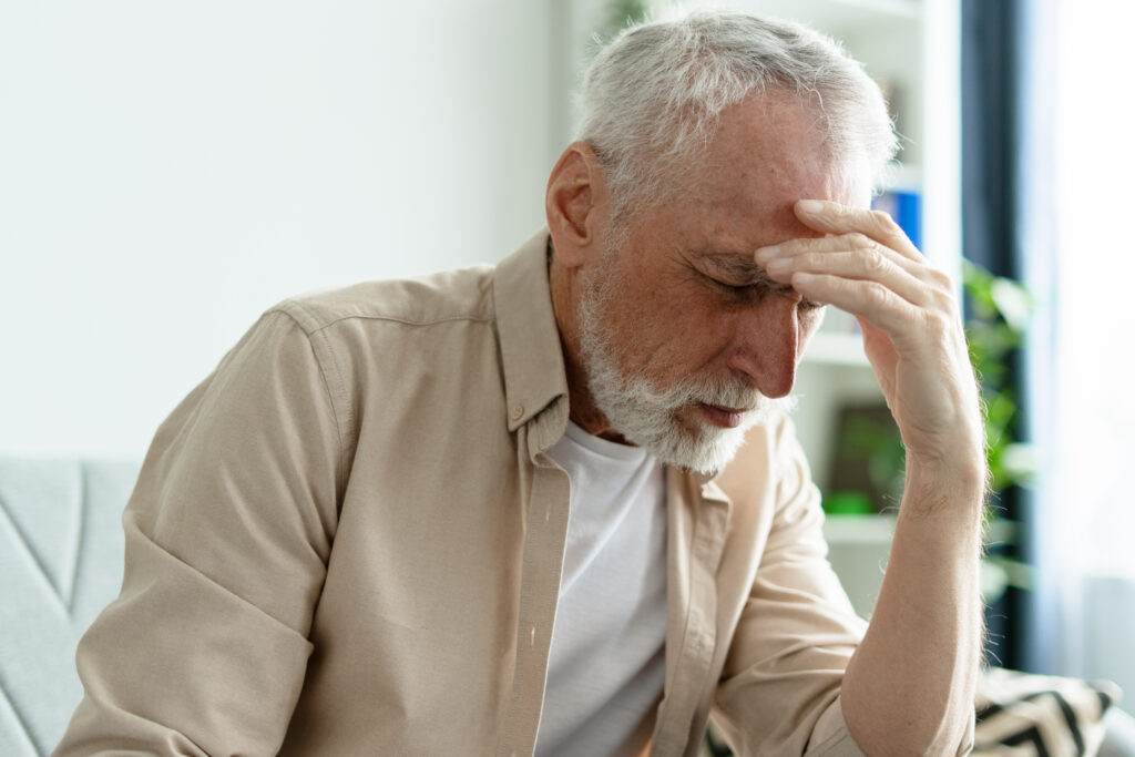 Elderly man holding his forehead while seated indoors, suggesting a headache, stress, or dizziness