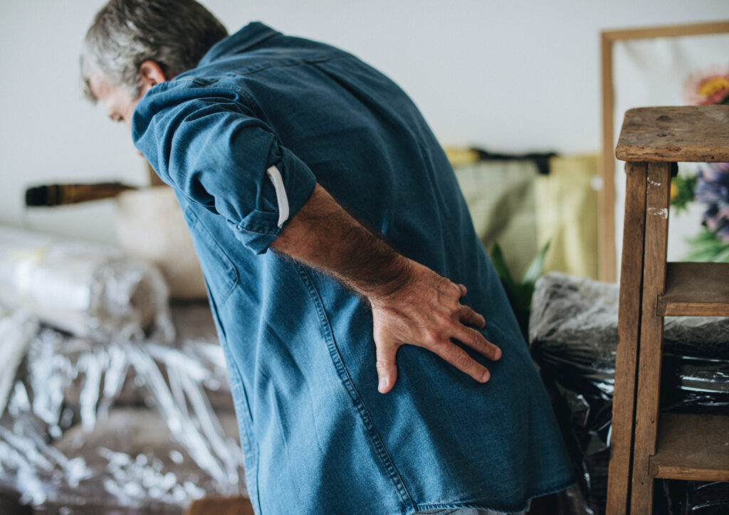Man holding his lower back in pain while standing indoors, suggesting back strain or chronic back discomfort
