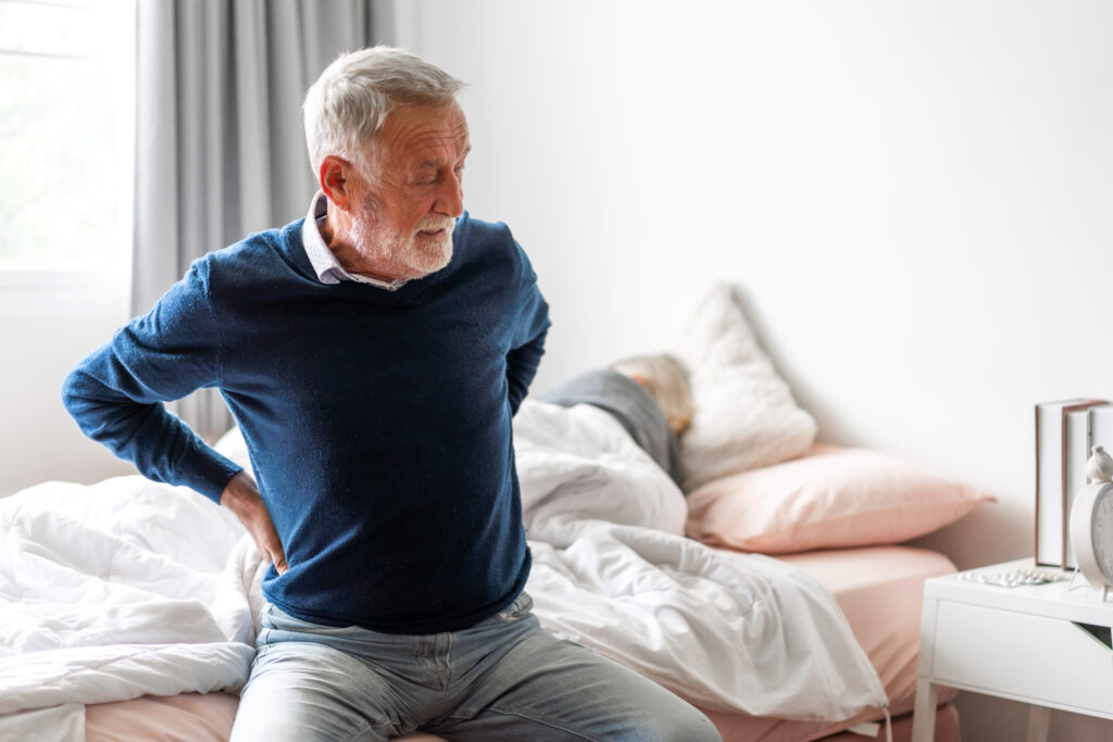Senior man sitting on a bed holding his lower back, showing signs of back pain or mobility discomfort