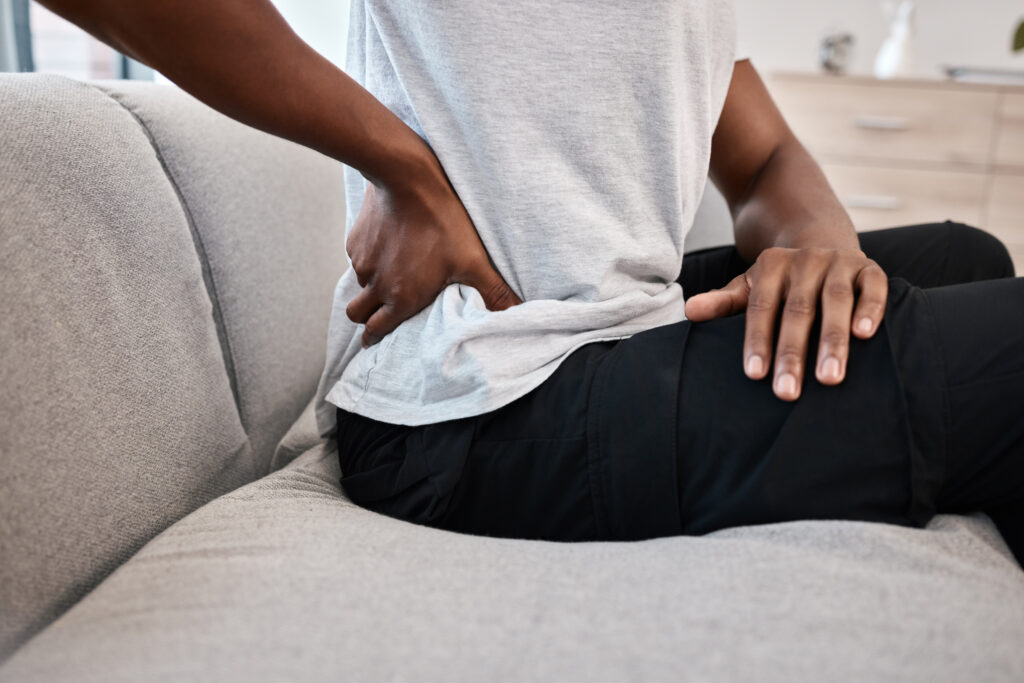 Person sitting on a couch holding their lower back, indicating back pain or muscle discomfort