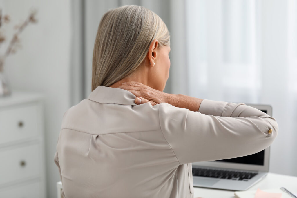 Woman touching the back of her neck while sitting at a desk, indicating neck pain or muscle tension